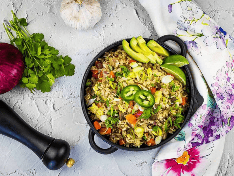 A cast-iron pan filled with Brisket Fried Rice, garnished with sliced avocado, lime wedges, jalape&ntilde;o slices, and fresh cilantro.