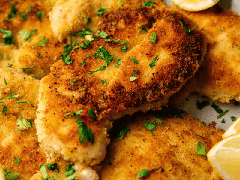 Breaded and pan-fried chicken cutlets garnished with chopped parsley, served with lemon wedges on the side.