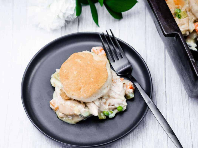 A black plate features a biscuit-topped serving of chicken pot pie, complete with chicken, peas, and carrots. A fork is placed on the plate, with a baking dish partially visible in the background.