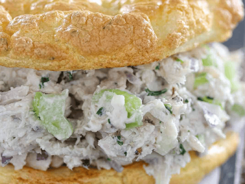 Close-up of a chicken salad sandwich with visible chunks of chicken, celery, and herbs, served in a fluffy, golden bread roll.