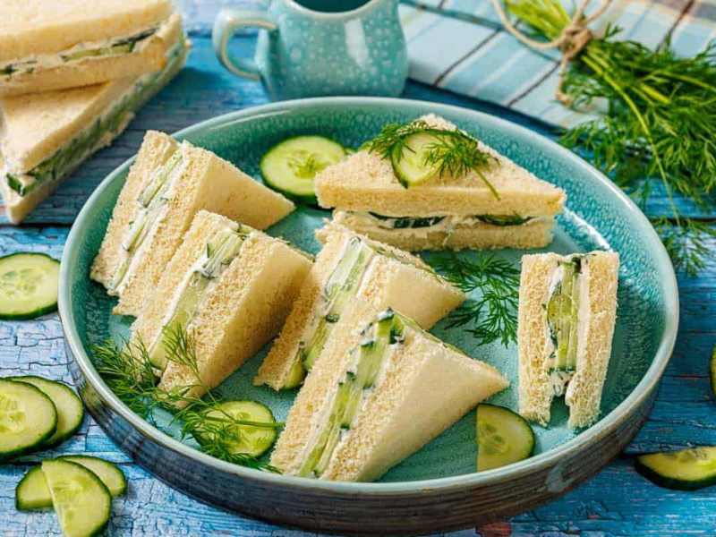 A plate of triangular cucumber sandwiches on white bread, garnished with fresh dill and cucumber slices, set on a blue wooden table.