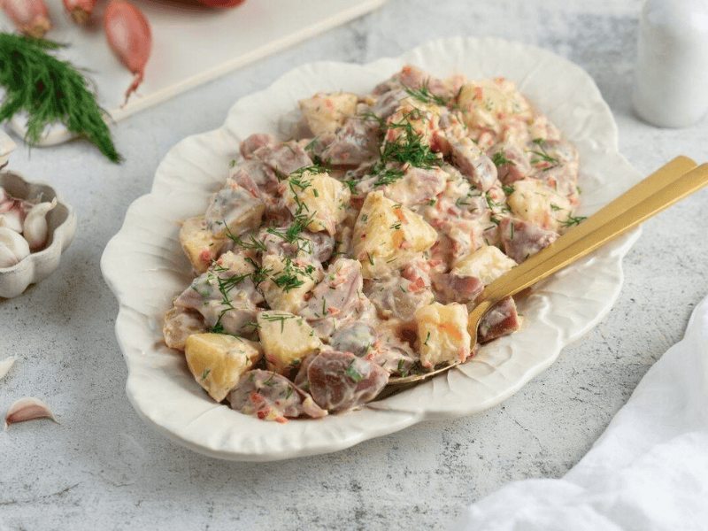 A serving dish filled with Classic Red Potato Salad with Pickles is garnished with fresh dill, with a gold-colored serving spoon resting in the dish.