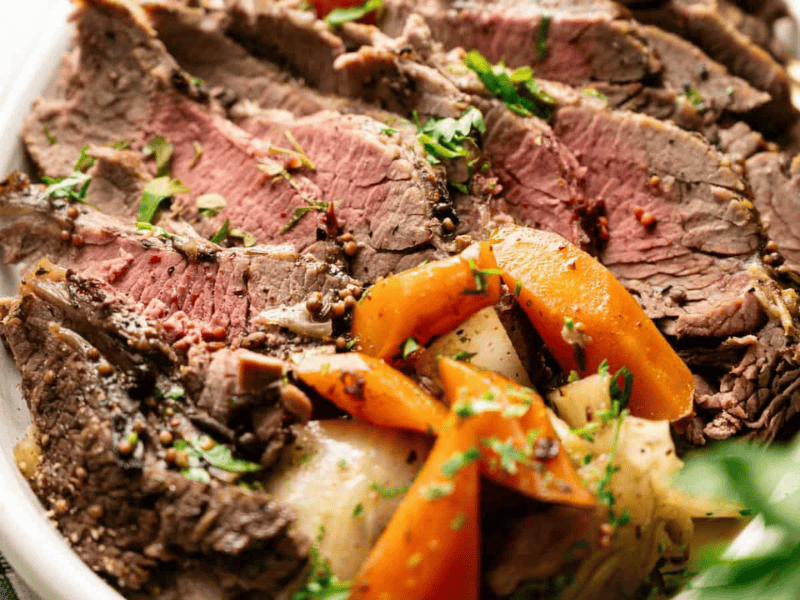 Sliced roast beef served on a plate with cooked carrots, potatoes, and herbs, placed on a checkered cloth.
