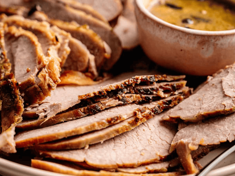 Sliced roast beef arranged on a plate with a bowl of gravy or sauce beside it.