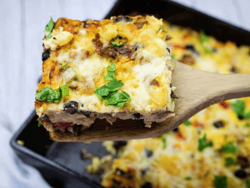 A wooden spatula holds up a square slice of baked casserole topped with melted cheese and chopped herbs, with more casserole in the background.