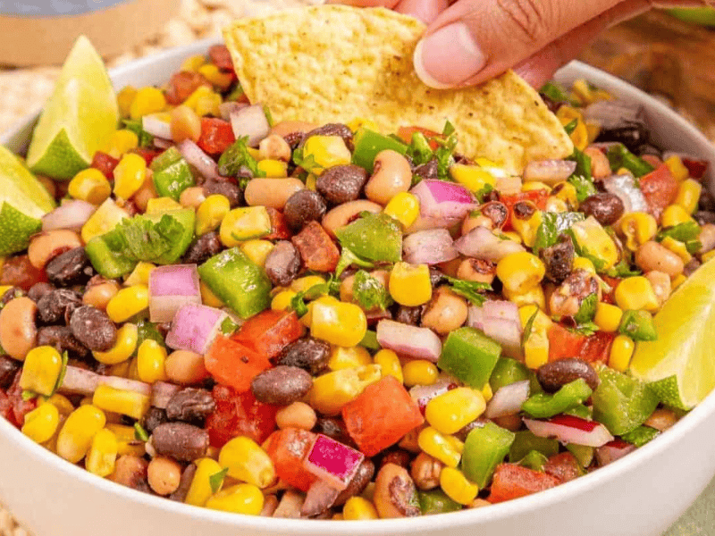 A bowl of Cowboy Caviar, garnished with lime wedges; a hand dips a tortilla chip into the salad.