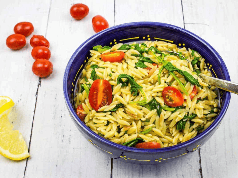 Creamy spinach orzo in a blue bowl with tomatoes nearby.