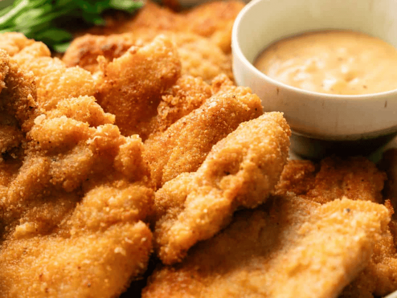 A plate of breaded and fried chicken cutlets served with lemon wedges, a bowl of dipping sauce, and greens in the background.