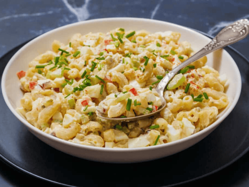 A bowl of Deviled Egg Pasta Salad with chopped vegetables and herbs, is set on a black plate, and a spoon rests in it.