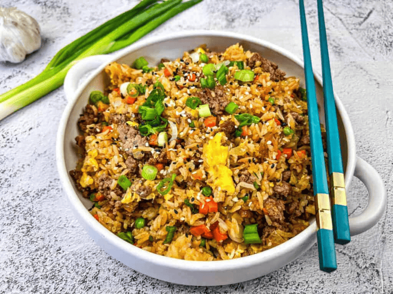A bowl of beef fried rice with scrambled eggs, diced vegetables, and chopped green onions, served with blue chopsticks on a light textured surface.