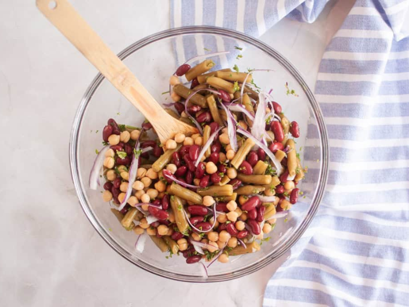 A glass bowl filled with a Three Bean Salad including kidney beans, chickpeas, green beans, and sliced red onion, with a wooden spoon and a striped cloth nearby.