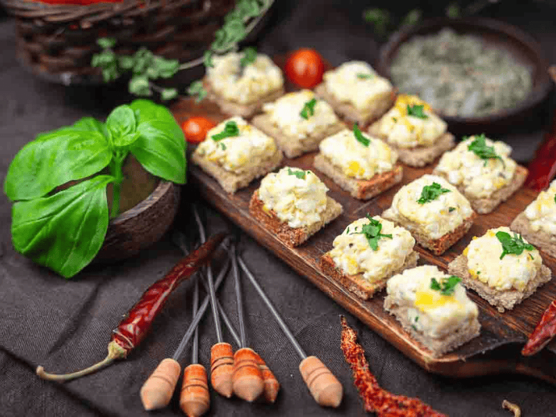 Several pieces of bread topped with a creamy feta and herb eggs, arranged on a wooden board, surrounded by fresh basil, dried chilies, and cherry tomatoes.
