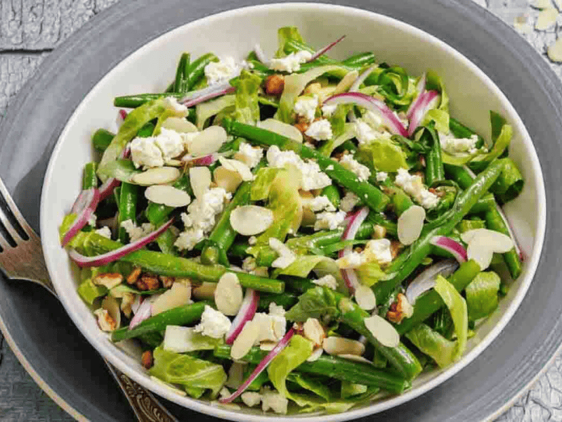 A bowl of green bean salad with sliced red onions, slivered almonds, goat cheese, lettuce, and walnuts, placed on a gray plate with a fork beside it.