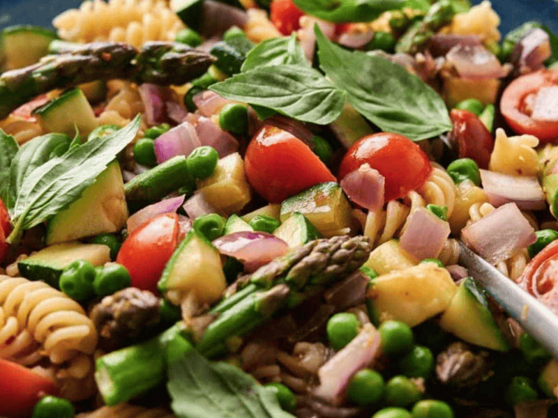 A colorful Pasta Primavera salad with cherry tomatoes, asparagus, peas, and spinach in a blue bowl.