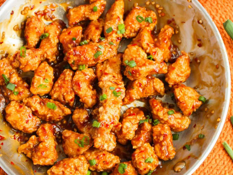 Stir-fried chicken pieces in a spicy sauce, garnished with chopped green onions, served in a round metal dish on an orange placemat.