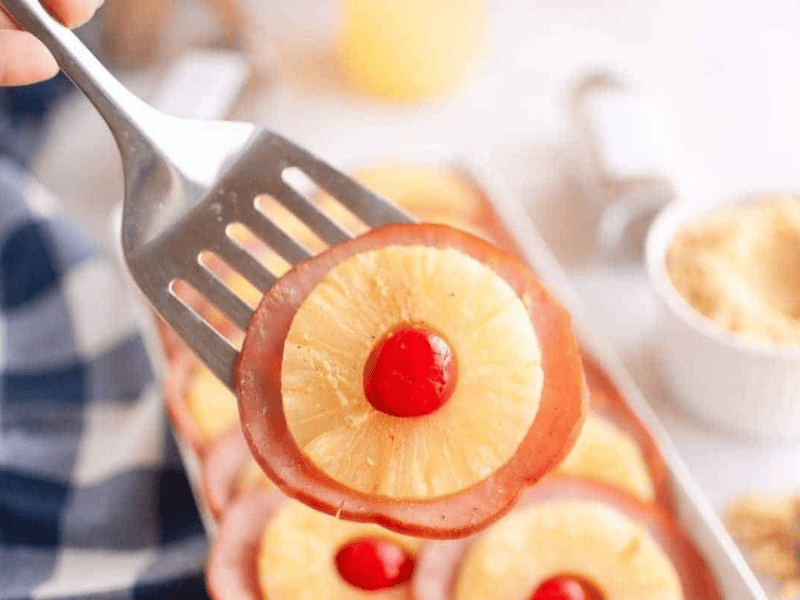 A metal spatula holds a slice of ham topped with a pineapple ring and a red maraschino cherry; more similar slices are visible in the background.