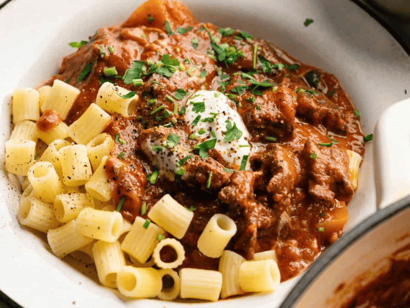 A plate of pasta with beef stew in tomato sauce, topped with a dollop of sour cream and chopped herbs.