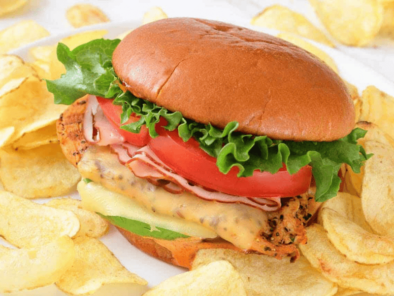 A grilled California chicken burger with lettuce, tomato, cheese, and ham on a bun, served with potato chips on a white plate.
