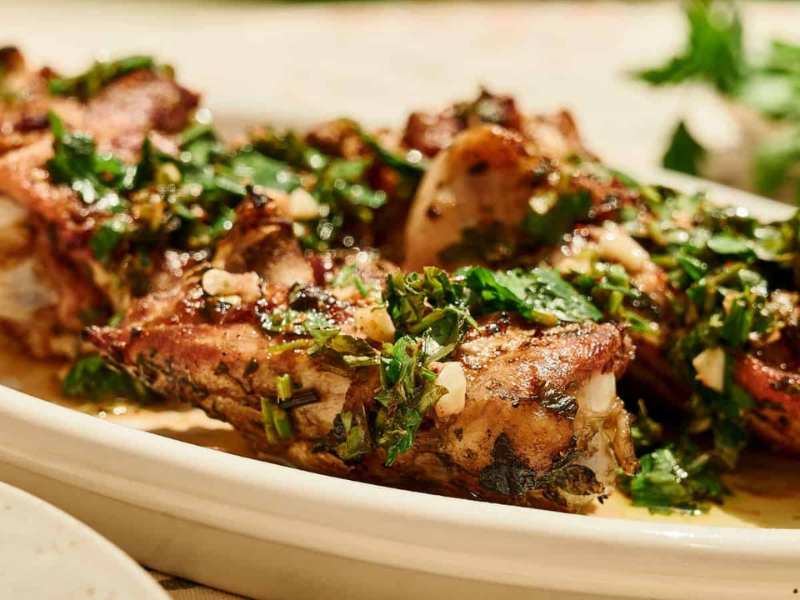 Grilled meat ribs garnished with chopped herbs and garlic, served on a white platter with fresh parsley in the background.