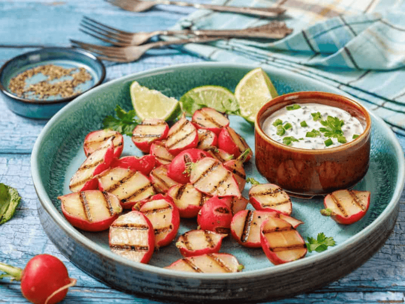 Grilled radish halves on a green plate with lime wedges and a bowl of herb dip; herbs and utensils in the background.