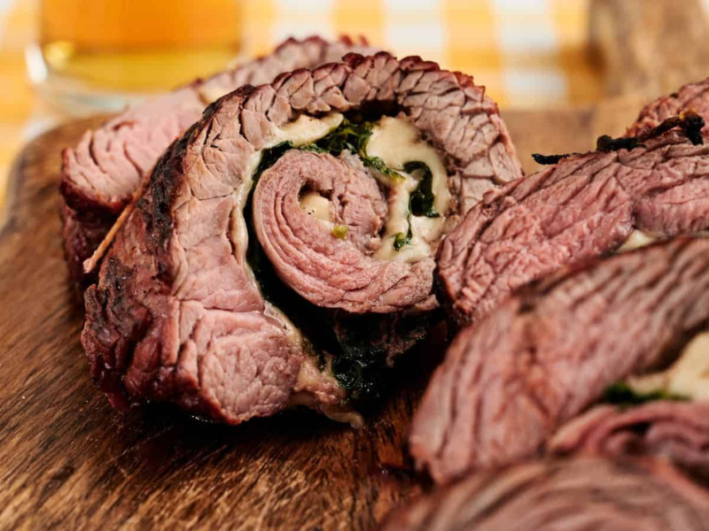 Close-up of sliced stuffed beef roulade with visible layers of meat, cheese, and spinach on a wooden board. A glass of beer is blurred in the background.