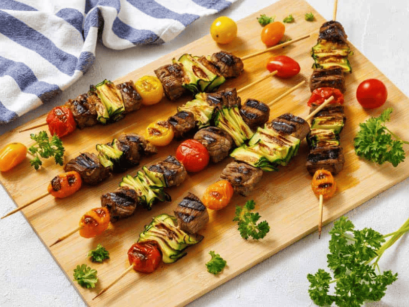 Grilled skewers with beef, zucchini, and cherry tomatoes on a wooden board, garnished with fresh parsley. A striped cloth and loose cherry tomatoes are nearby.