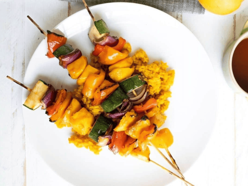 Three Grilled Teriyaki Mango Skewers with bell peppers, zucchini, and onions served on a bed of yellow rice on a white plate.