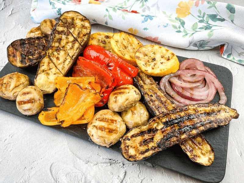 Grilled vegetables including zucchini, mushrooms, red onions, peppers, and squash, arranged on a rectangular black platter.
