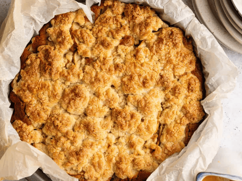 A baked apple crumble sits in a round, parchment-lined pan next to a stack of plates and spoons on a light countertop.