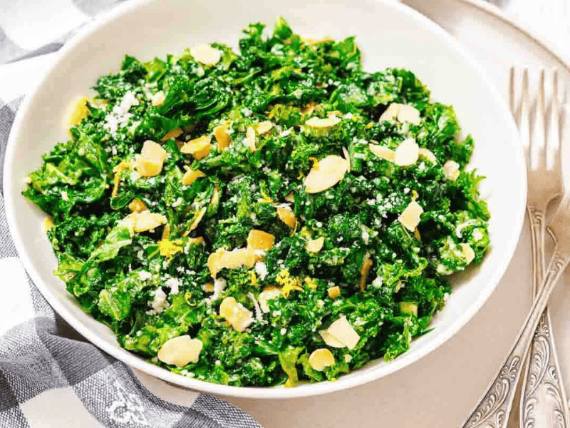 A white bowl filled with chopped kale salad topped with sliced almonds and grated cheese, placed on a plate beside two forks and a checkered napkin.