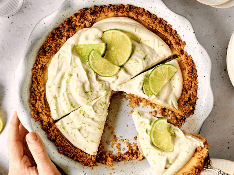 Key lime pie with a slice removed on a white plate. Topped with lime wedge and zest, the crust is golden brown. A hand touches the plate edge, and a small bowl with cream is nearby.