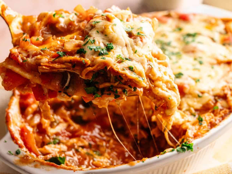 A serving of cheesy baked lasagna is being lifted from a casserole dish, showing layers of pasta, sauce, and melted cheese with parsley sprinkled on top.