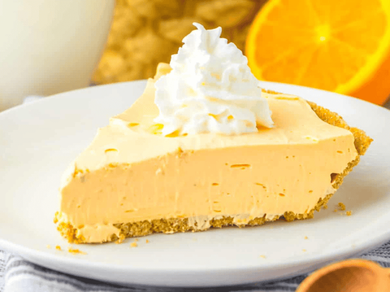 A slice of cream pie on a white plate, topped with whipped cream. An orange slice and a drink are in the background.