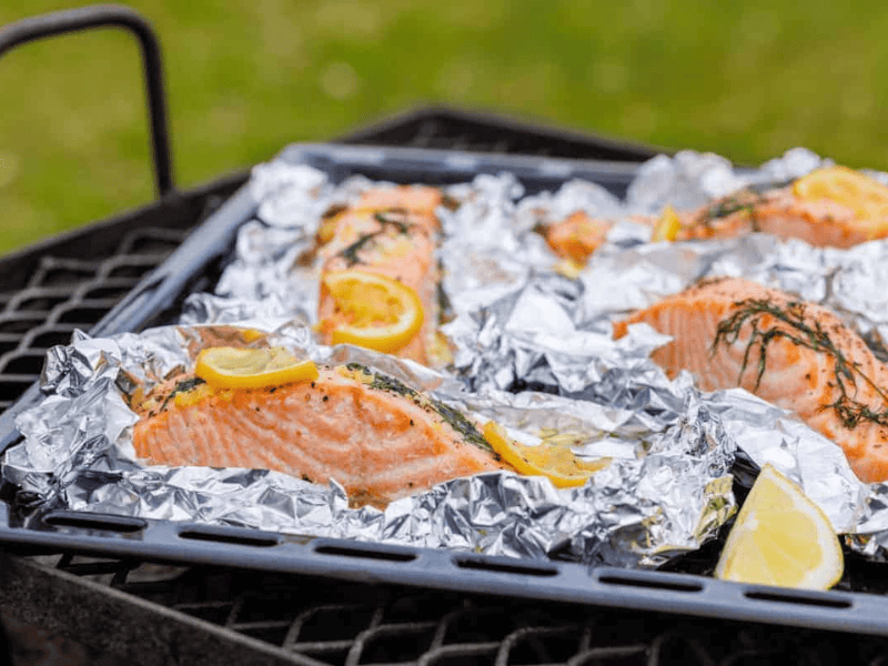 Four servings of lemon-dill salmon in foil packets, with lemon slices and dill on top, arranged on a black grill grate with a green grass background.
