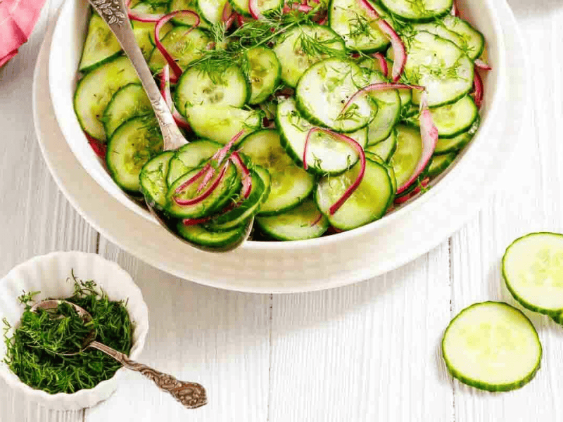 A bowl of cucumber salad with sliced red onions and fresh dill, next to a small dish of chopped dill and cucumber slices on a white wooden surface.
