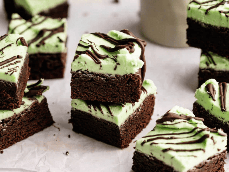 Brownie squares topped with a layer of green mint frosting and drizzled with chocolate, arranged on parchment paper.