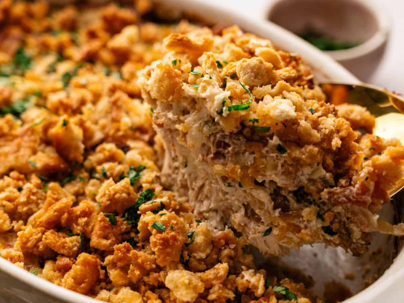 A serving spoon lifts a portion of a baked casserole topped with golden, crispy breadcrumbs and garnished with chopped herbs from a white dish.