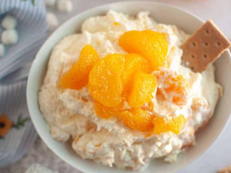 Overhead shot of a white bowl filled with creamy Orange Fluff Salad, topped with mandarin oranges and served with graham crackers.