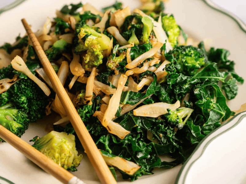 A plate of stir-fried kale, broccoli, and sliced onions, served with a pair of chopsticks on the side.