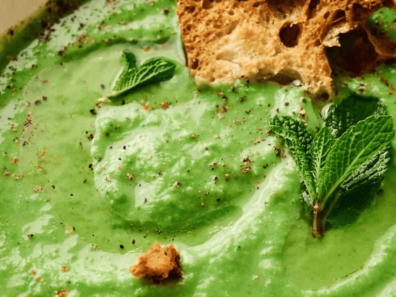 A bowl of creamy green pea soup garnished with mint leaves, black pepper, and pieces of crusty bread.