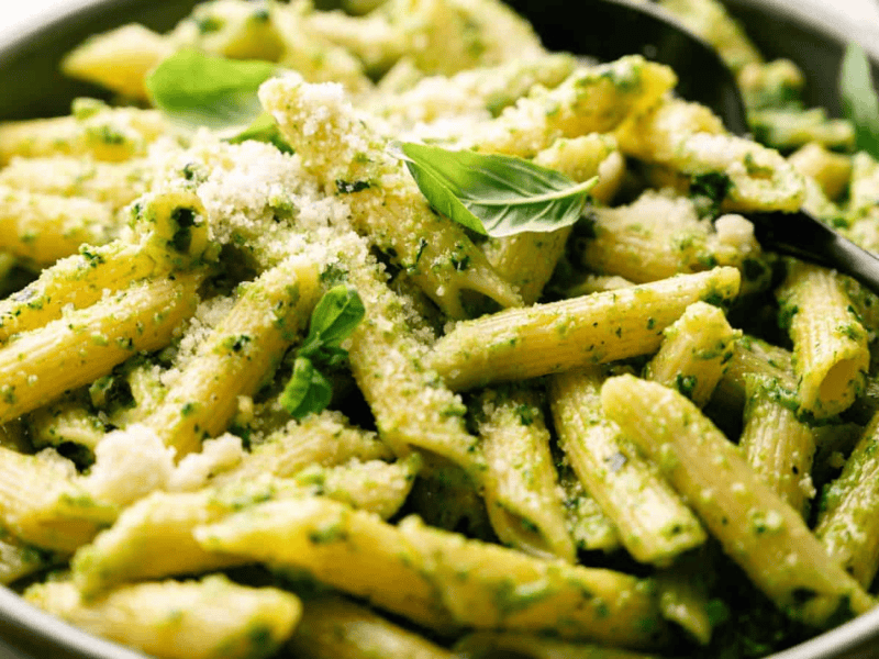 A bowl of penne pasta with green pesto sauce, topped with grated cheese and fresh basil leaves, with a black serving spoon.