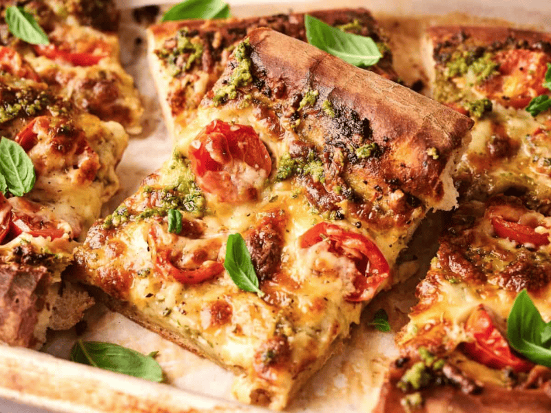 Rectangular slices of pizza topped with melted cheese, tomato, green pesto, and fresh basil leaves on a baking tray.