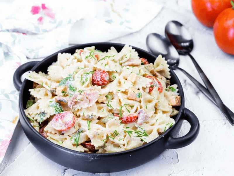 A black bowl filled with picnic pasta salad with chopped vegetables and herbs, placed on a white surface next to spoons and fresh tomatoes.