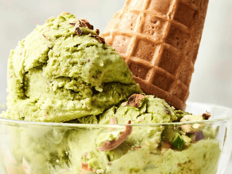 A glass bowl holds two scoops of pistachio ice cream topped with crushed pistachios, with a waffle cone placed upside down in the bowl.