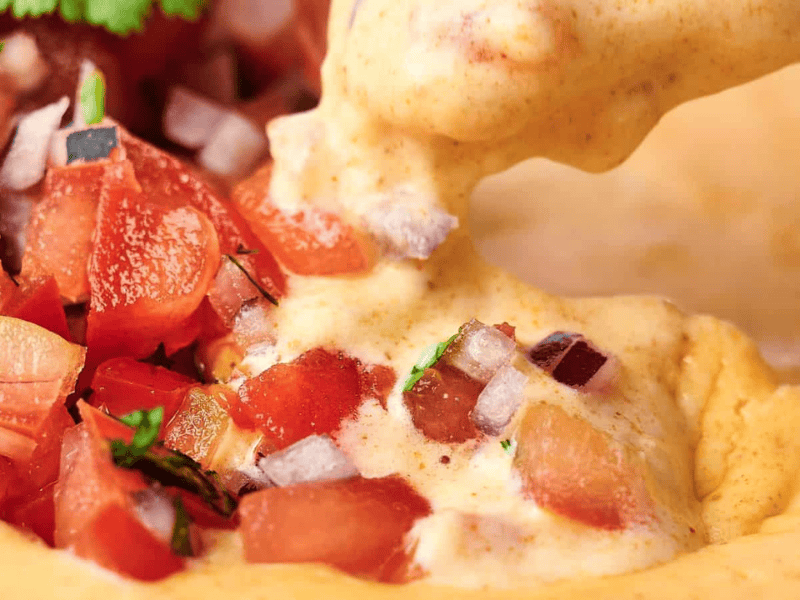 Close-up of creamy cheese dip being poured over chopped tomato salsa with diced onions and cilantro.