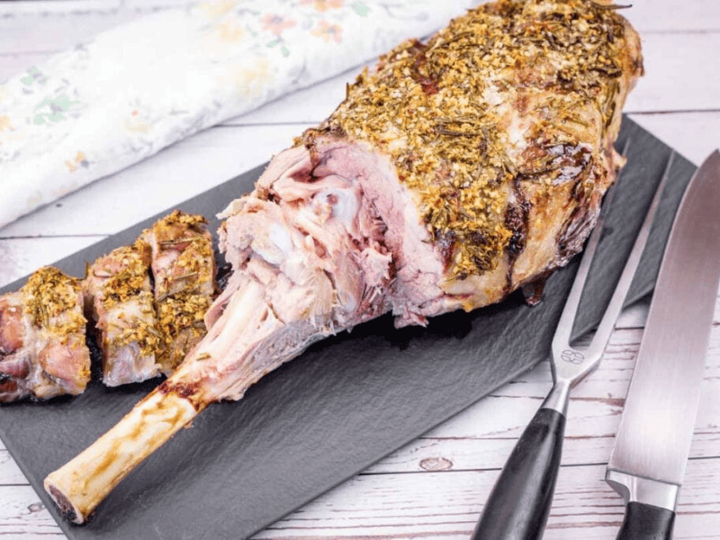 Roast leg of lamb with herb crust, partially sliced, displayed on a slate board with a carving fork and knife beside it.