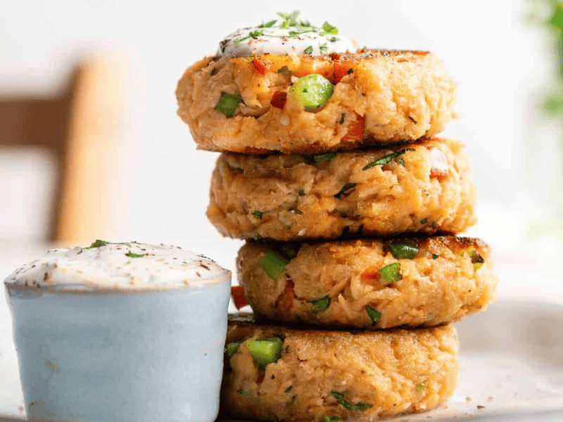A stack of four vegetable patties is plated next to a small cup of creamy dipping sauce, garnished with herbs.