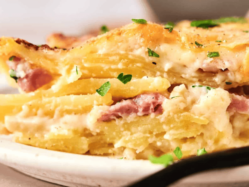 A close-up of a slice of potato casserole with layers of potatoes, ham, and creamy sauce, garnished with chopped parsley on a plate.
