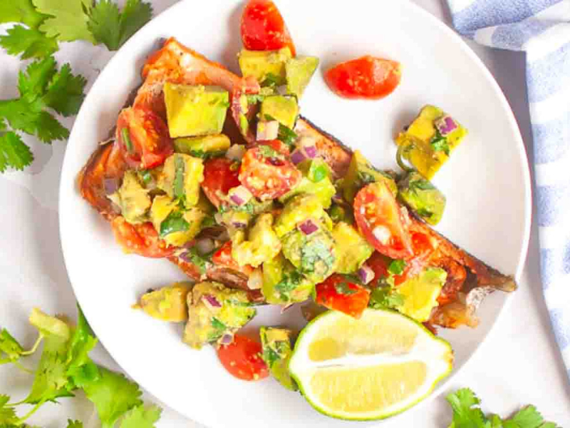 Grilled salmon topped with avocado, tomato, and onion salsa, served with a lime wedge on a white plate.