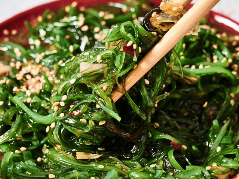 A close-up of seaweed salad with sesame seeds on a red plate, with chopsticks picking up a portion.